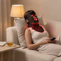 Woman relaxing on a sofa wearing the black and gold LED face and neck mask glowing red